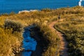 Field of grass by the river with blue waters and warm light during sunset with a man walking on a path walk. Royalty Free Stock Photo