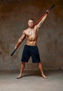 A fighter or karate man, stands in a fighting artificially pose and holds a bamboo stick on gray wall studio background.