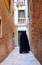 figure in a hood walks through a narrow city alley wearing a worn black tabard as a cloak