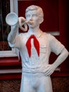 The figure of a pioneer with clarion in his hand near building. White sculpture of a boy with a red tie from the time of the