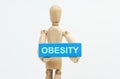 A figurine of a man holds in his hands a blue wooden block with the inscription OBESITY. The figurine is out of focus