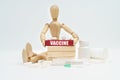 A figurine of a man sitting among pills lifts a red wooden block with an inscription VACCINE from a wall of blocks.