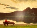 Film effect. Alone man sits leg over on bench beside an azure mountain lake. Man relax and watch high peaks of Alps above lake mir Royalty Free Stock Photo