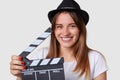 Film making concept. Pleasant looking joyful woman wears hat, has broad smile, holds film slate or clapper board, being in good mo Royalty Free Stock Photo