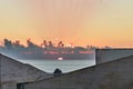 The final glimpse of the sun dips below the ocean horizon, casting soft rays through clouds and illuminating modern rooftops at Royalty Free Stock Photo