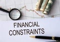 FINANCIAL CONSTRAINTS - words on a white sheet against the background of a chart, magnifying glass and banknotes