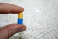 Fingers holding blue and yellow bright capsule pill, with blur a lot of white drugs tablets on background