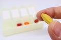 Fingers Holding a Yellow Supplement Pill with Blurry Pill Organizer Case in Background