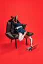 Finishing tasks. Businessman lying on armchair in weird pose, working on laptop against red studio background Royalty Free Stock Photo