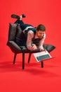 Finishing tasks. Businessman lying on armchair in weird pose, working on laptop against red studio background Royalty Free Stock Photo