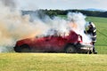 A firefighter extinguishes a car fire. Red car on fire, smoke and firefighter. Accident at the meadow