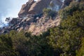 Firefighters climb rocks and use hoses in the middle of a brush fire on a rocky hillside