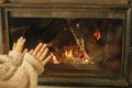 Fireplace heating in winter, alternative to gas and electricity. Woman in cozy sweater warming up hands at fireplace in rustic