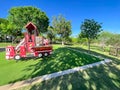 Firetruck themed playground with green artificial grass turf carpet surrounding by tall mature trees under clear blue sky in