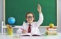 First day at school. A happy schoolgirl with glasses smiling raised her hand up while sitting at a table in the Royalty Free Stock Photo