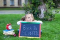 First-grader girl sitting on green grass with chalkboard, alarm clock and books near school. Back to school. Education concept. Cu Royalty Free Stock Photo