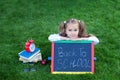 First-grader girl sitting on green grass with chalkboard, alarm clock and books near school. Back to school. Education concept. Cu Royalty Free Stock Photo