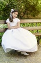 First Holy Communion, little girl in a white communion dress sitting on a bench in a park with plants. Sunbathing photography Royalty Free Stock Photo