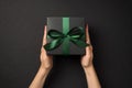 First person top view photo of female hands holding black giftbox with green satin ribbon bow on  black background Royalty Free Stock Photo