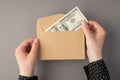 First person top view photo of female hands holding open craft paper envelope with hundred dollars banknote on grey