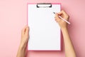 First person top view photo of female hands holding pink pen and pink clipboard with paper sheet on isolated pastel pink Royalty Free Stock Photo