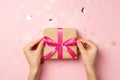 First person top view photo of female hands tying pink ribbon bow on craft paper giftbox over large shiny sequins on isolated