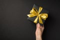 First person top view photo of hand giving black giftbox with golden satin ribbon bow on  black background with empty Royalty Free Stock Photo