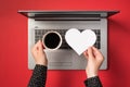 First person top view photo of hands holding cup of coffee and white paper heart over open grey laptop on isolated red background Royalty Free Stock Photo