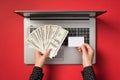 First person top view photo of hands holding fan of dollars and small rectangular white card over open laptop on  red Royalty Free Stock Photo