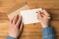 First person top view photo of hands holding pen over white card and craft paper envelope on isolated wooden table background with Royalty Free Stock Photo