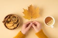 First person top view photo of hands in pullover holding yellow maple leaf cup of tea with lemon wooden bowl with cookies dried