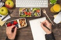 First person top view photo of hands taking blueberry from lunchbox healthy food and writing in notepad apples glass of juice