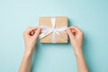 First person top view photo of hands tying white satin ribbon bow on craft paper giftbox on isolated pastel blue background Royalty Free Stock Photo