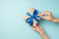 First person top view photo of hands unpacking craft paper giftbox with vivid blue satin ribbon bow on isolated pastel blue Royalty Free Stock Photo