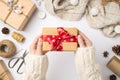 First person top view photo of woman`s hands in cozy sweater holding craft paper giftbox with red ribbon bow over scarf christmas Royalty Free Stock Photo