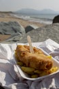 Fish and chips, battered cod, in a tray wrapped in paper with a wooden fork on a beach. Traditional British food Royalty Free Stock Photo