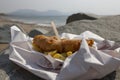 Fish and chips, battered cod, in a tray wrapped in paper with a wooden fork on a beach Royalty Free Stock Photo