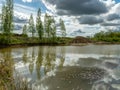 A fish pond, in the water of which there are colorful cumulus clouds and trees, there are recently dug gravel piles on the shore