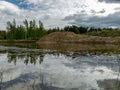 A fish pond, in the water of which there are colorful cumulus clouds and trees, there are recently dug gravel piles on the shore