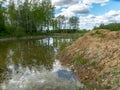 A fish pond, in the water of which there are colorful cumulus clouds and trees, there are recently dug gravel piles on the shore