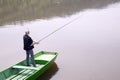 Fisherman Casting A Fishing Rod From The Green Boat On The Lake And Patiently Waiting For Fish To Take A Bait
