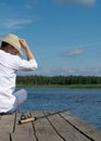 A fisherman with a hat sits on the pier of the river and looks at the fishing rod on a hot sunny day Royalty Free Stock Photo