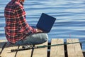 A fisherman man works on a laptop, sits on a wooden pier near the lake