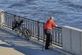 A fisherman in an orange T-shirt on the pier with a fishing rod next to the bike. A pier with a handrail by the river