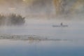 Fisherman in a punt boat fishing with rods, thick fog covers still surface of a river, bulrush, cane and willows grow on a bank Royalty Free Stock Photo