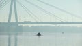 Fisherman sailing under big bridge on inflatable boat, misty weather, big city