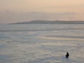 A fisherman sits near the ice hole on the frozen river on a beautiful sunset on a frosty day