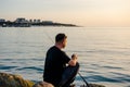 A fisherman sits on a rock by the sea with a fishing rod at sunset. Single sea fishing. Day at the beach. Copy space for text