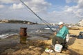 Fisherman sitting on a chair on the rocks by the sea while fishing with fishing stick on a sunny morning in Sliema , Malta