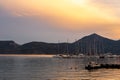 Fishing and sailing boats docked in Adamas Port on Milos Island, Greece during golden hour sunset, with calm flat sea